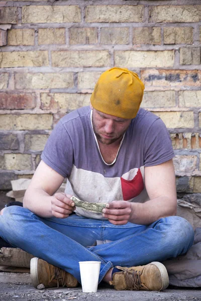 いくつかのお金でホームレスの男性は悲しいことに壁の近くに座っています。お金問題はないです。 — ストック写真
