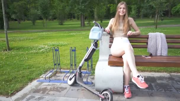 幸せな笑顔の若いブルネットの女性は電動スクーターの近くのベンチに座ってカメラを見ています 安定したショット — ストック動画
