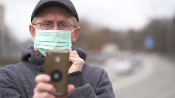ウイルス問題チームだ 医療用マスクのシニア白人男性が街の通りに立って スマートフォンを見ている 肖像画 — ストック動画