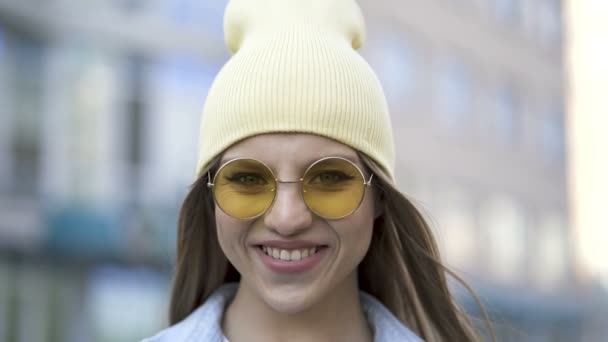 4Kだ 黄色の帽子と眼鏡の本当の若い笑顔の女の子は 都市エリアに立っています ストリートポートレート — ストック動画