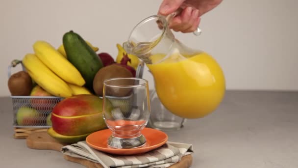 用玻璃杯从酒瓶中倒入果汁 一杯果汁 黄色令人神清气爽的夏季饮料 新鲜的芒果汁橙汁桌上的果篮 厨房里新鲜的水果和果汁 维生素饮料 — 图库视频影像