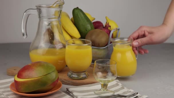 用玻璃杯从酒瓶中倒入果汁 一杯果汁 黄色令人神清气爽的夏季饮料 新鲜的芒果汁橙汁桌上的果篮 厨房里新鲜的水果和果汁 维生素饮料 — 图库视频影像