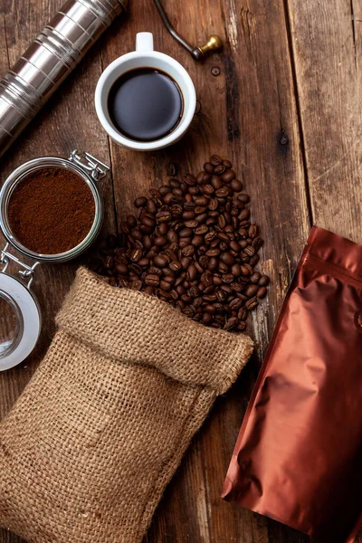 Tahta bir masada bir poşet kahve. Kahve çekirdekleri ve bir kavanozda öğütülmüş. Elle kahve öğütücü. Üst görünüm, metin için yer. Kahverengi arka plan. Pakette kahve paketliyorum. İçkili bir fincan..