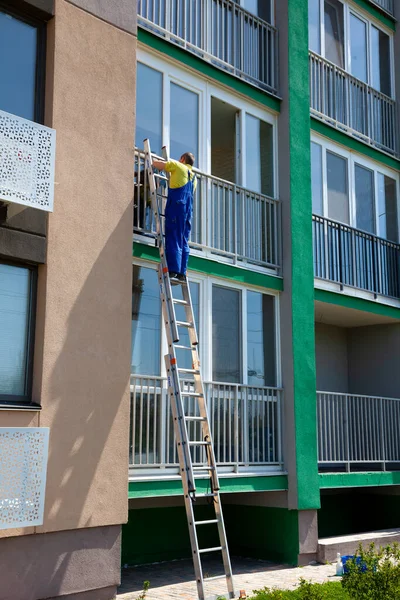 A man washes windows on the street standing on the stairs. Cleaning company. Worker in blue overalls washes a window of a multi-storey apartment building.