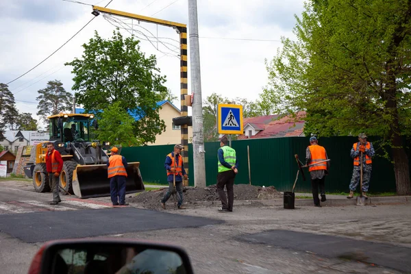 Bucha, Ukrayna - 18 Mayıs 2020: Yol işçileri. Sokakta yol onarımı. Asfaltı asfaltlamak ve yollara çukur açmak. Parlak yelekli adamlar sokakta çalışır. Açık hava işi. Traktör ve yolda kürekle dolaşan adam..