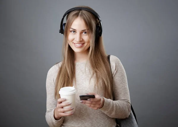 Attractive young smiling girl in headphones is holding a phone and a cup of coffee.
