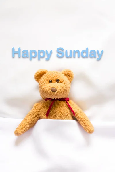Teddybär liegt im weißen Bett mit der Botschaft "Happy Sunday"" — Stockfoto
