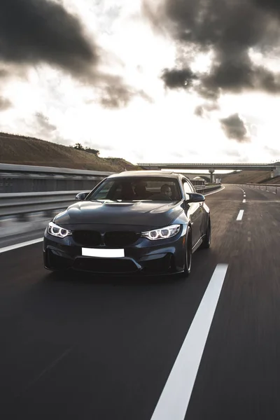 Siyah sedan kara bulutların altında yolda gidiyor.