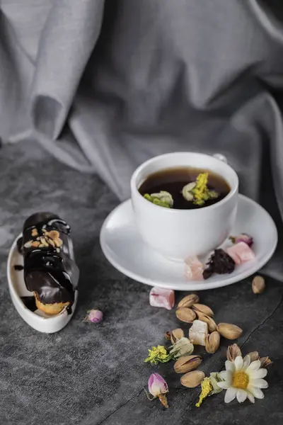 Чашка чая с фисташками и шоколадным эклером, украшенная цветами — стоковое фото