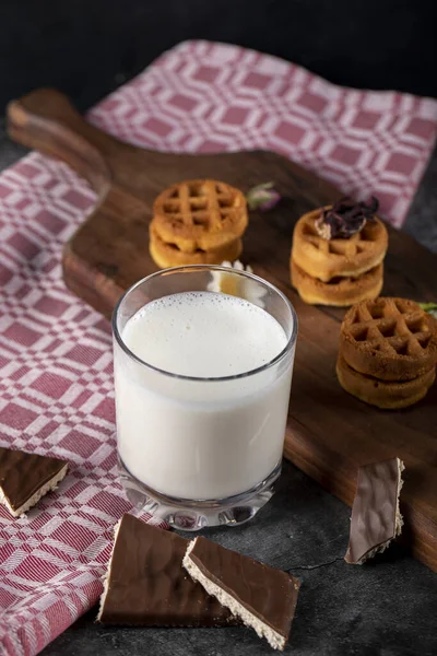 Pequenos waffles em uma placa de madeira com um copo de leite em um pedaço de toalha vermelha — Fotografia de Stock