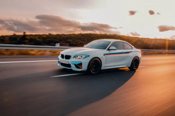 Beyaz sedan güneş ışığının altında otoyolda ilerliyor.