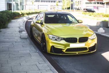 Merkez yolun kenarına park edilmiş sarı spor araba, küçük yeşil bölgeler.