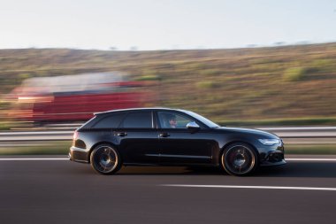 Gün batımında otoyolda siyah spor arabaları diğer arabaların yanından geçiyor.