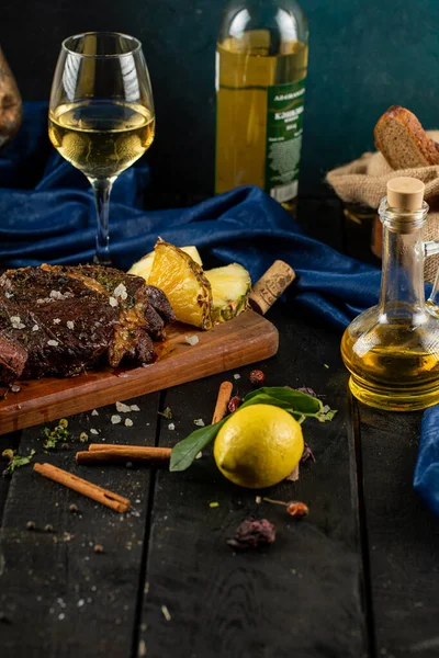 Barbacoa de carne en rodajas servida sobre una mesa de madera, rodajas de piña, copa de vino, limón y canela alrededor sobre una mesa negra — Foto de Stock