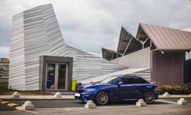 Mavi spor sedan panoramik bir binaya park edilmiş.