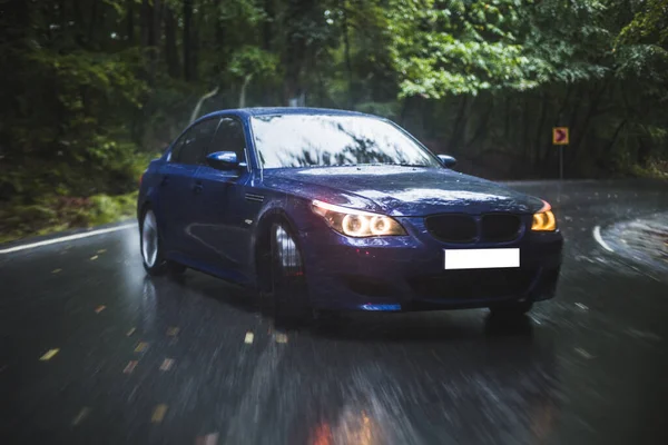 Conducir coche azul, luces amarillas encendidas en el bosque oscuro en la lluvia ducha, aparcamiento en el medio de la carretera — Foto de Stock