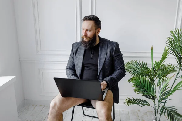 Cara barbudo em uma jaqueta sem calças funciona em um laptop em um quarto brilhante . Fotografia De Stock