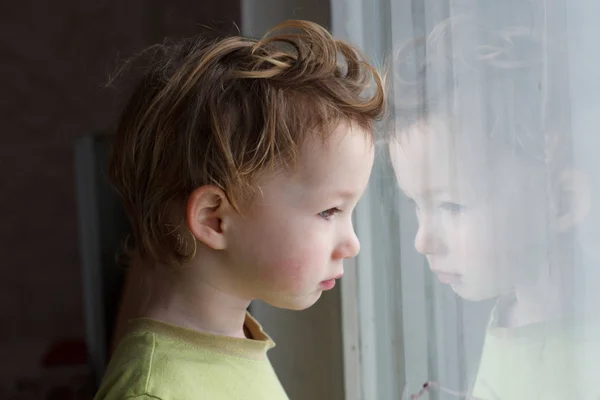 Malý chlapec sedí u okna a myslet na něco. Má krásné velké vlasy. Roztomilé dítě. — Stock fotografie