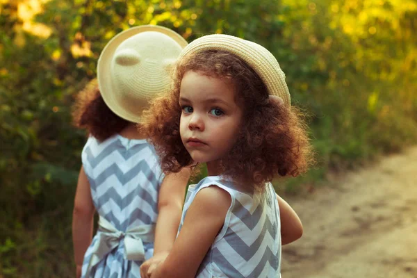 Retrato de una niña rizada triste y su hermana gemela. Un sombrero en la cabeza. Retrato de cerca al aire libre. Chica se dio la vuelta —  Fotos de Stock