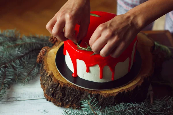Holly Şubeler ve kestane ile eski Beyaz ahşap masa üzerinde rozmarine ve kırmızı meyveler ile kadının el tarafından işlem ev yapımı Noel kırmızı ganache pasta dekorasyon. Karanlık rustik tarzı.