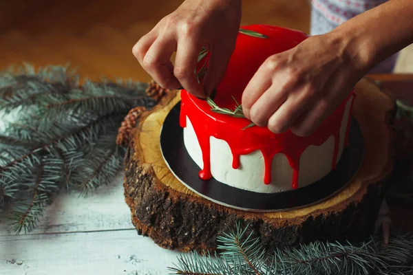 Holly Şubeler ve kestane ile eski Beyaz ahşap masa üzerinde rozmarine ve kırmızı meyveler ile kadının el tarafından işlem ev yapımı Noel kırmızı ganache pasta dekorasyon. Karanlık rustik tarzı.