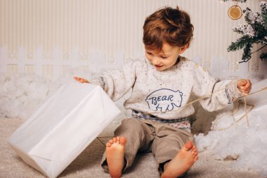 Sevimli küçük çocuk Noel ağacı yakınındaki bir hediye ile oturuyor. Yeni yıl hediyesi ile mutlu bir çocuktur. Beyaz sarışın yürümeye başlayan çocuk