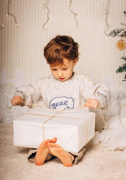Menino bonito está sentado com um presente perto da árvore de Natal. A criança está feliz com o presente de Ano Novo. Caucasiano loira criança menino — Fotografia de Stock
