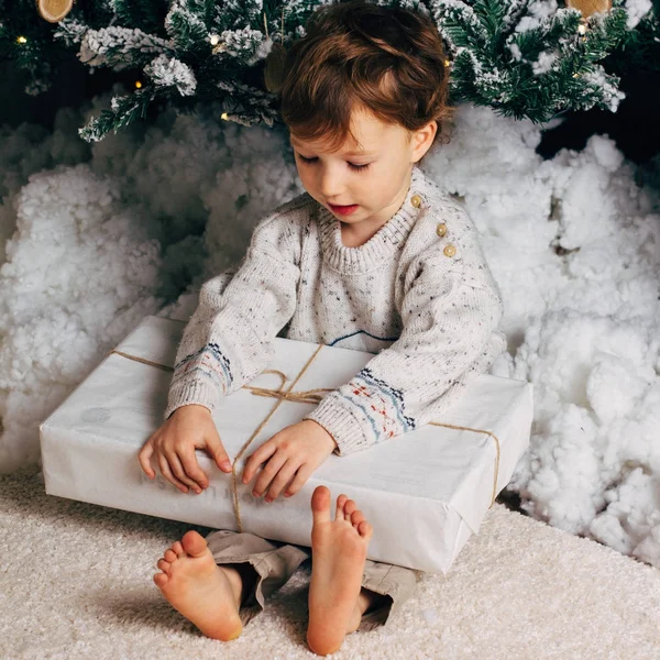 Bonito menino sentado com uma caixa de presentes ao redor da árvore de Natal. Criança branca loira adorável. Foto quadrada — Fotografia de Stock