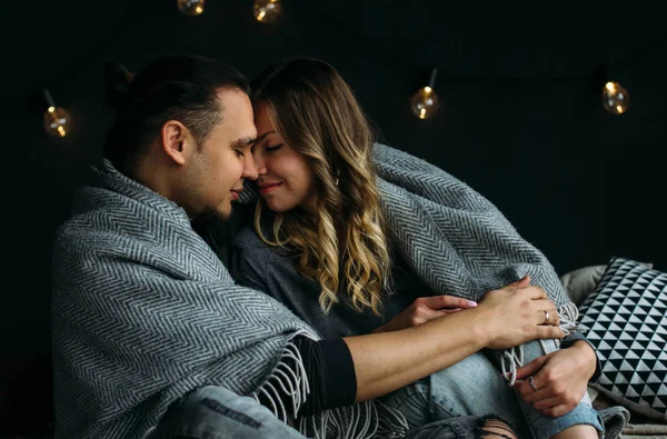 Boyfriend and girfriend touching each other sitting on bed with grey blankets and pillows. Cozy room with lights, romantic and love concept. Couple kissing and smiling — Stock Photo, Image