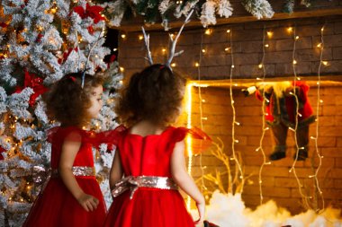 Yakınındaki güzel Noel ağacı Noel şömine bakarak iki kıvırcık küçük kız. İkizler kırmızı elbiseler Santa şömine görünümlü. Sevimli geyik boynuzu hoop kafasına çocuklarla