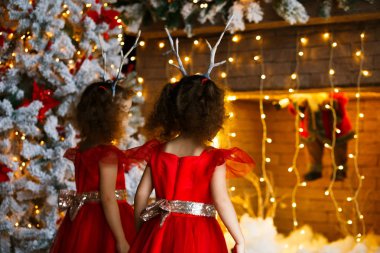 Yakınındaki güzel Noel ağacı Noel şömine bakarak iki kıvırcık küçük kız. İkizler kırmızı elbiseler Santa şömine görünümlü. Sevimli geyik boynuzu hoop kafasına çocuklarla