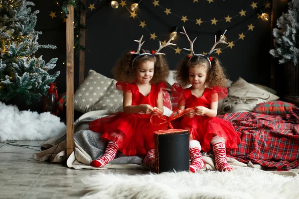 Crianças Abertura Presente de Natal. Duas garotinhas com chifres de veado na cabeça. Gêmeos em vestidos vermelhos sentados na cama no quarto acolhedor . — Fotografia de Stock