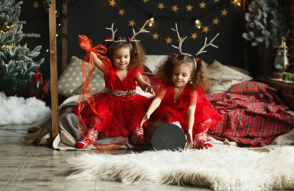 Dzieci, otwierając prezent. Dwa małe todder dziewczyna z jelenie rogi na głowie. Bliźniaki w czerwone sukienki siedział na łóżku w przytulny pokój. — Zdjęcie stockowe