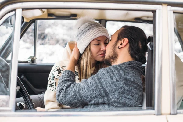 A Valentine couple sitting inside a snow retro car and kissing in a forest. Romantic Beauty girl in a hat and handsome man in a grey pullover. Horisontal picture — Stock Photo, Image