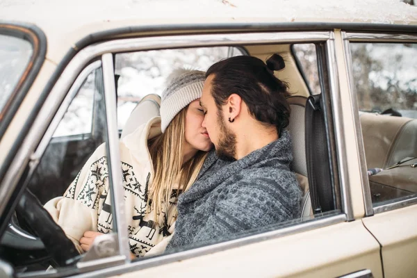 Valentine Młoda siedzi wewnątrz auta retro śniegu i Całowanie w lesie. Romantyczne piękno dziewczyna w kapeluszu i przystojny mężczyzna w szary sweter. Obraz poziomy — Zdjęcie stockowe