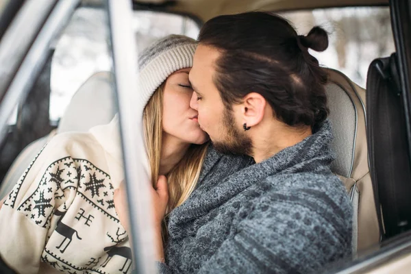 A Valentine couple sitting inside a snow retro car and kissing in a forest. Romantic Beauty girl in a hat and handsome man in a grey pullover. Horisontal picture — Stock Photo, Image