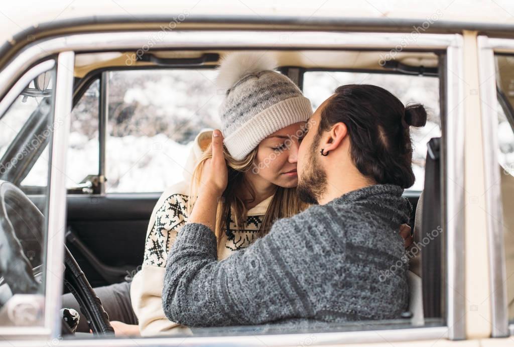 A Valentine couple sitting inside a snow retro car and kissing in a forest. Romantic Beauty girl in a hat and handsome man in a grey pullover. Horisontal picture
