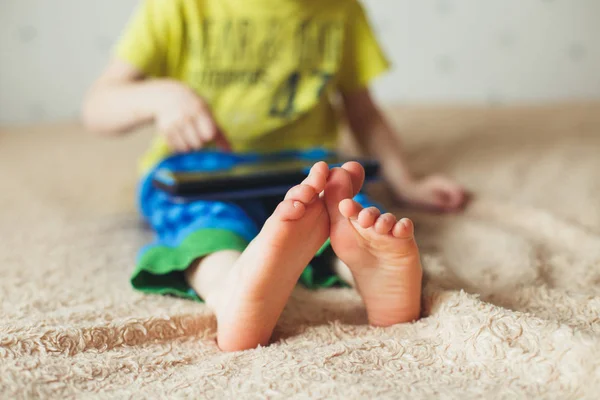 Kind mit Tablet auf dem Bett sitzend und Cartoons guckend. selektiver Fokus auf die Füße. Netter Junge in grünem T-Shirt und blauer Hose — Stockfoto