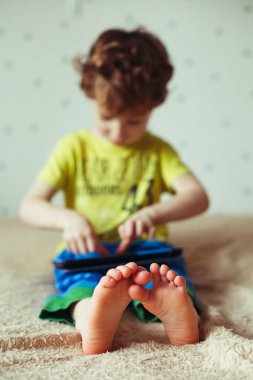 Yatağın üstüne oturan ve çizgi film izlerken tablet ile evlat. Seçici odak ayakları üzerinde. Yeşil t-shirt ve Mavi pantolon şirin çocuk. Dikey fotoğraf