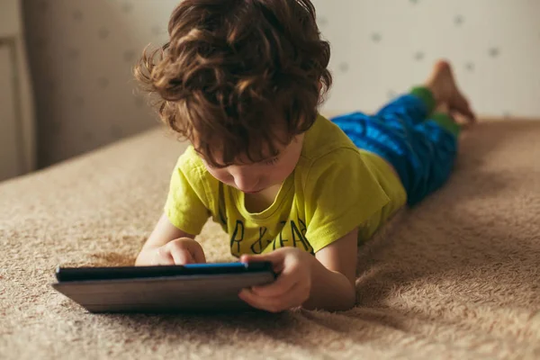 Милый маленький мальчик, использующий блокнот. Ребёнок играет с цифровым планшетом, лежащим на кровати. Свободное время. Технология и интернет-концепция — стоковое фото