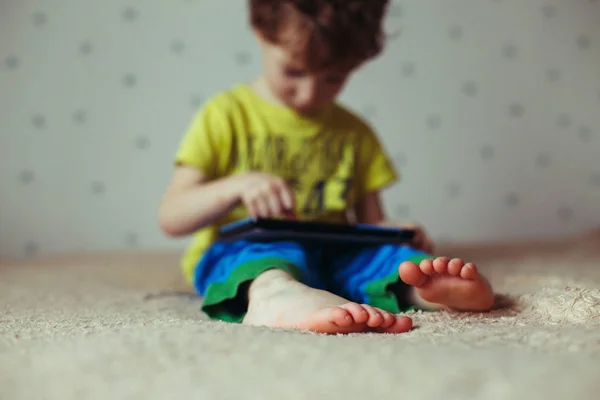Niño con la tableta sentado en la cama y viendo dibujos animados. Enfoque selectivo en los pies. Lindo chico en camiseta verde y pantalones azules — Foto de Stock