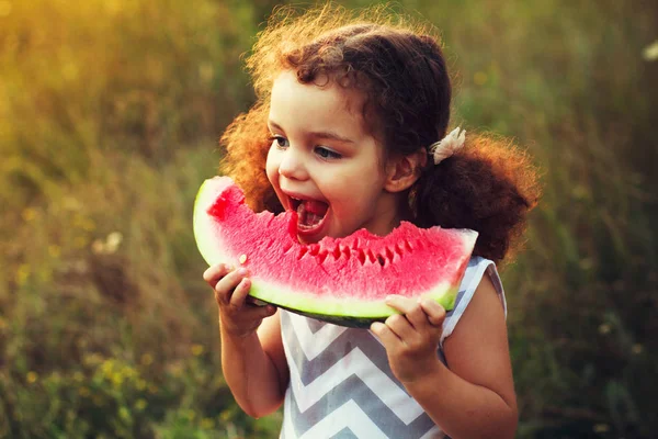 Komik karpuz, sağlıklı meyve snack, sevimli yürümeye başlayan çocuk kıvırcık saçlı bir sıcak yaz günü güneşli bir bahçede oynarken yeme bir inanılmaz güzel kıvırcık saçlı küçük kız portresi
