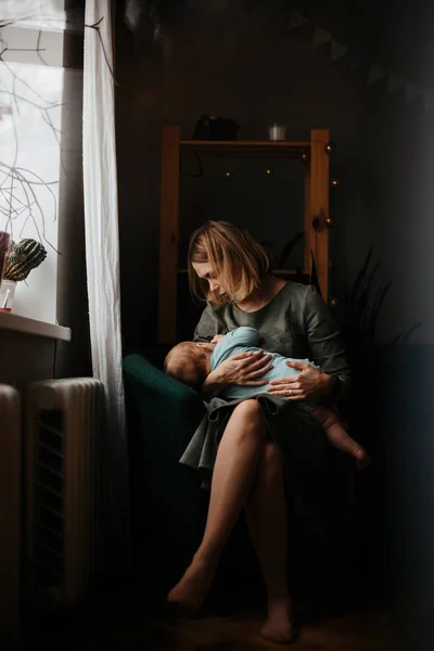 Madre y bebé en casa. Lactancia materna y nutrición infantil saludable. Mujer joven amamantando a su hijo recién nacido sentada en mecedora blanca en una guardería soleada con cuna y ventana. Padres e hijos en casa — Foto de Stock