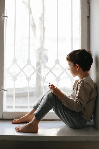 Child in home quarantine looking out of the window with his smartphone, protection against viruses during coronavirus and flu outbreak. Children and illness COVID-2019 disease concept — Stockfoto