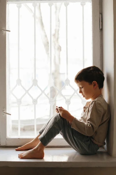 Niño en cuarentena domiciliaria mirando por la ventana con su smartphone, protección contra virus durante el coronavirus y el brote de gripe. Niños y enfermedades COVID-2019 concepto de enfermedad —  Fotos de Stock