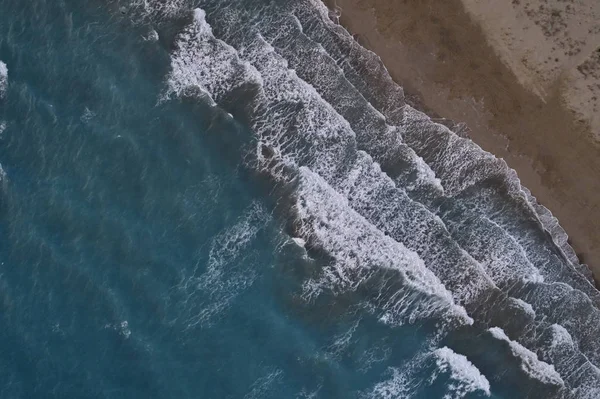 White foamy sea waves splash to deserted sandy beach in the evening. Evening surf. Aerial top view from UAV drone