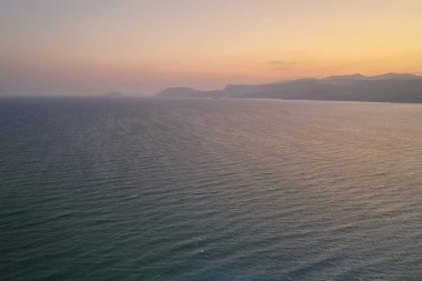 Ön planda deniz dalgaları ve ufukta sisli dağlar. Akşam sörfü