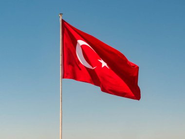 Arkaplanda açık mavi gökyüzüne karşı bayrak direğinin tepesinde Türk bayrağı sallanıyor