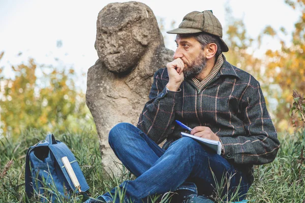 Learned historian sits before stone sculpture on mound — Stock Photo, Image