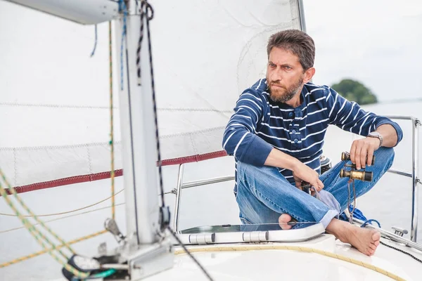 Man sits on bow of sailing yacht and holds binoculars — Stock Photo, Image